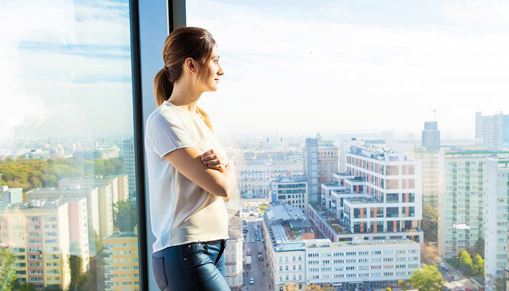 Bild einer Frau, die aus einer großen Fensterfläche auf ein Stadtpanorama blickt